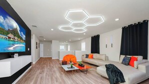 A modern living room with a large flat-screen TV, a sofa, a coffee table, and a unique hexagonal ceiling light fixture. - 6BR at Windsor Cay - Private Pool, Spa & Themed Rooms (Clermont)