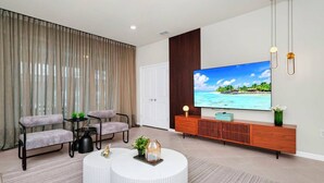 A modern living room with a large flat-screen TV, two grey upholstered chairs, a white coffee table, and a wooden entertainment unit. - 6BR at Windsor Cay - Private Pool, Spa & Themed Rooms (Clermont)