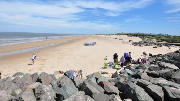 Ein Strand mit felsigem Ufer, entspannten Personen und blauem Himmel.