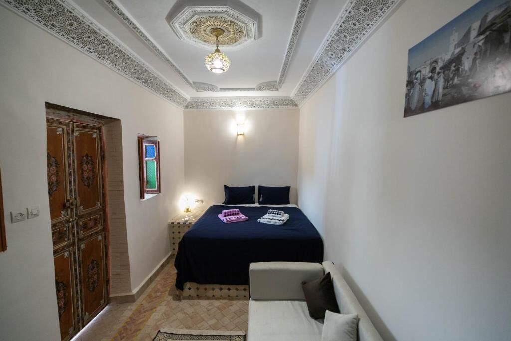 A bedroom with a bed, a chair, a door, a wall-mounted light, and a ceiling with intricate patterns.