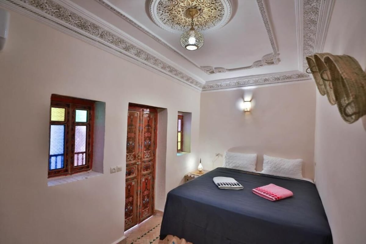 A bedroom with a bed, a nightstand, a lamp, a decorative ceiling, and a window with a view.