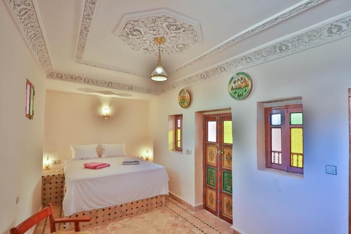 A room with a bed, a chair, a decorative ceiling, and a door with colorful glass panels.