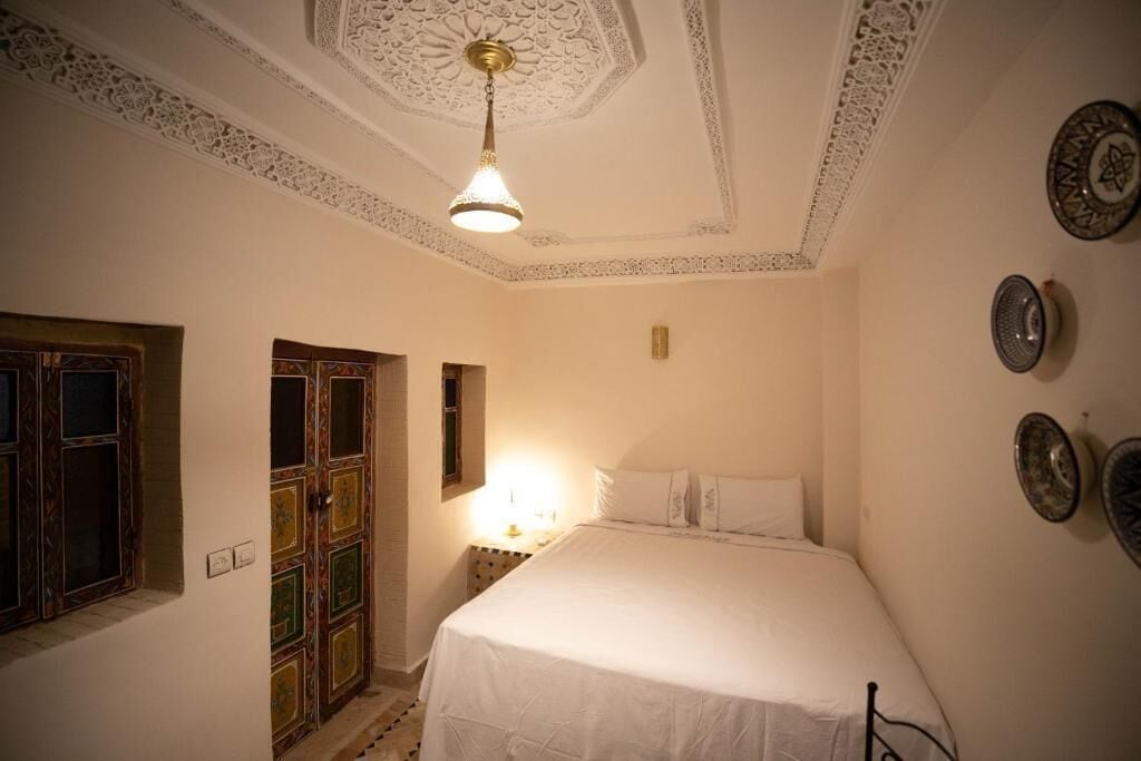 A bedroom with a bed, a nightstand, a wall-mounted lamp, a decorative ceiling, and a door with intricate patterns.