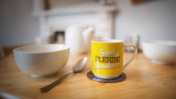 Une tasse jaune avec l’inscription « Bonjour, rayon de soleil » posée sur un dessous de verre, un bol blanc et une cuillère sur une table en bois.