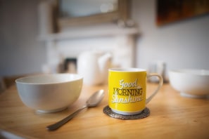 Une tasse jaune avec l’inscription « Bonjour, rayon de soleil » posée sur un dessous de verre, un bol blanc et une cuillère sur une table en bois.