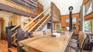 A spacious dining area with a long wooden table and cushioned chairs, a staircase with wooden railings, and a large window with a view of the outdoors.