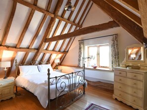 Ein Schlafzimmer mit geneigter Decke, einem Bett, einer Kommode, einem Fenster mit Vorhängen und einer Lampe.