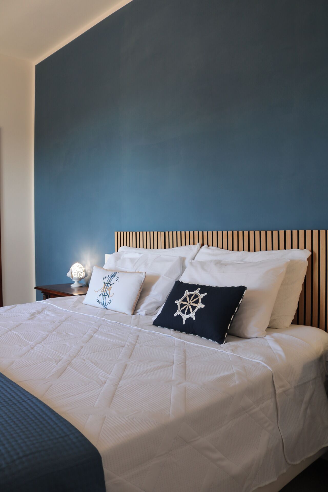 Un letto rifatto con cura, lenzuola bianche e due cuscini decorativi, una testiera in legno e un comodino con una lampada.