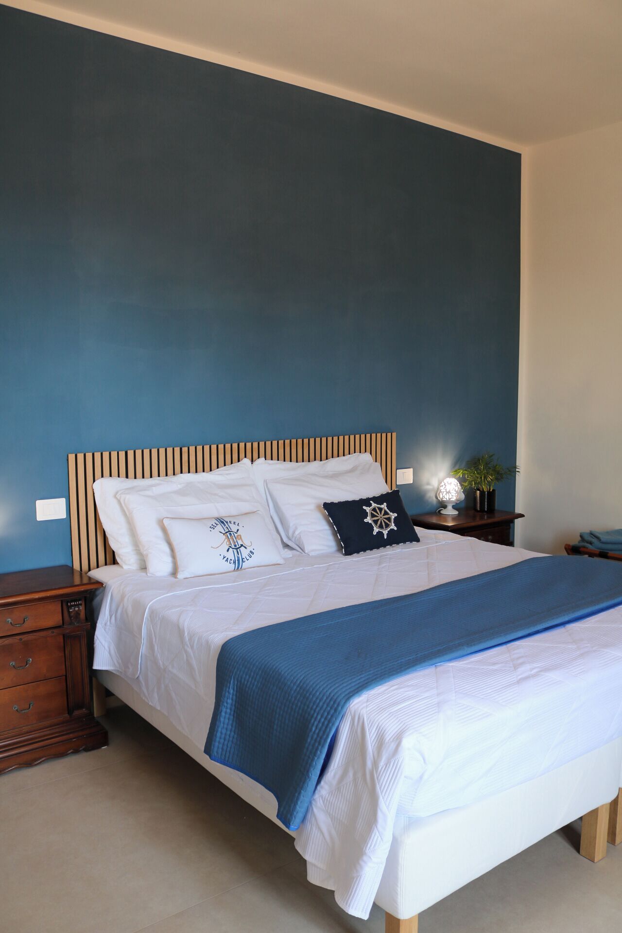 Un letto ben rifatto con una coperta blu, una testiera in legno e due comodini con lampada e pianta.