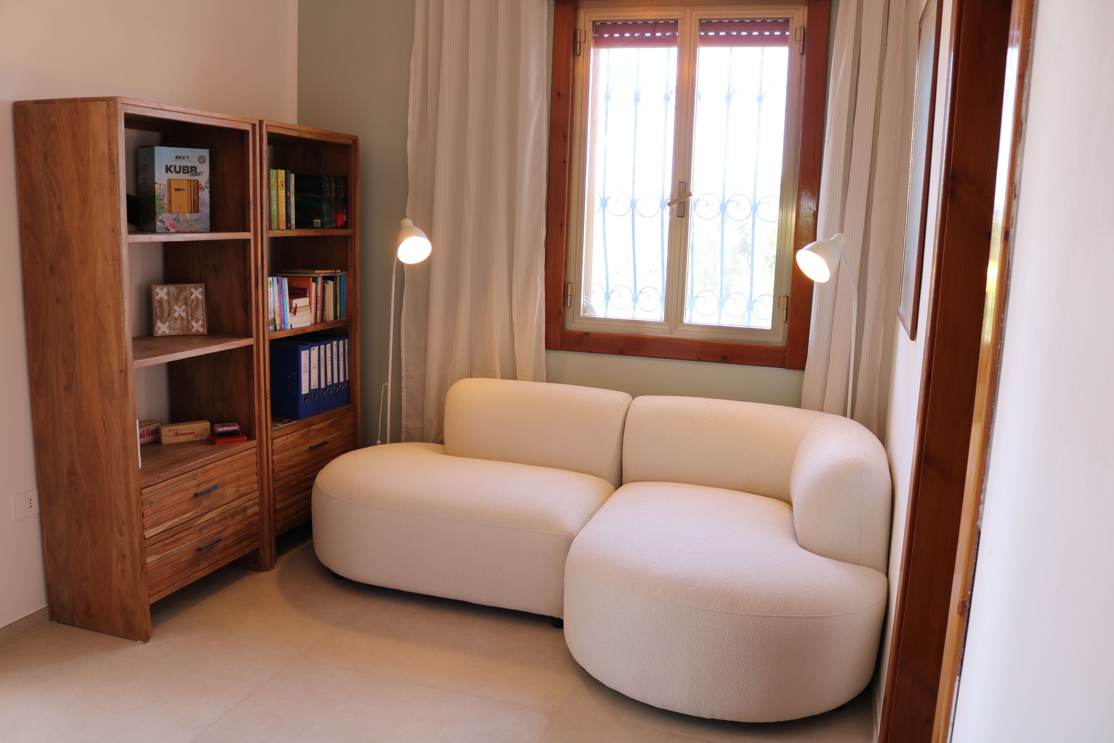 Una stanza con una libreria in legno, un divano beige, una finestra con tende e due lampade a parete.