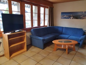 Een woonkamer met een televisie, een blauwe bank, een klein rond tafeltje en een raam met gordijnen.