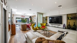 A modern living room with a sofa, chairs, a TV, and a dining area with a table and chairs. - Design-Led 3-Bed Home with Garden, Sauna, Hot Tub & Parking (Greater London)