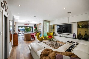Ein modernes Wohnzimmer mit einer Couch, Sesseln, einem Fernseher und einem Essbereich mit Tisch und Stühlen.