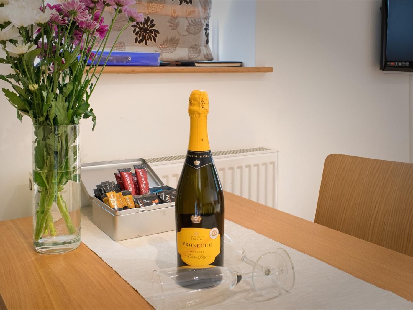 A bottle of Prosecco on a table with a glass, a vase of flowers, and a box of chocolates.