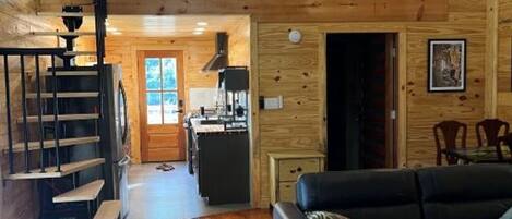 A wooden cabin interior with a loft bedroom, a dining area, and a living room with a black leather sofa.