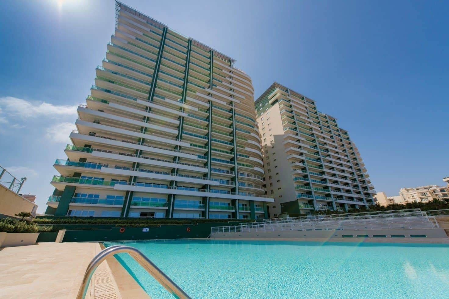 A high-rise hotel with multiple balconies, a swimming pool, and a clear blue sky.