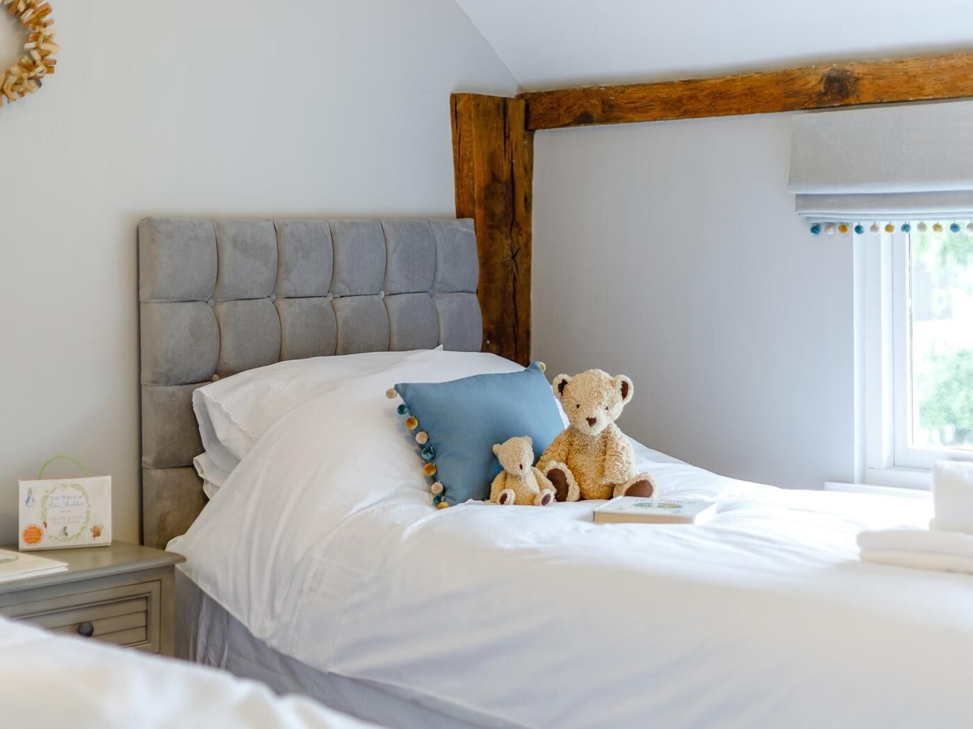 A bedroom with a tufted headboard, a bed with white linens, two pillows, a teddy bear, and a bedside table with a book and a decorative item.