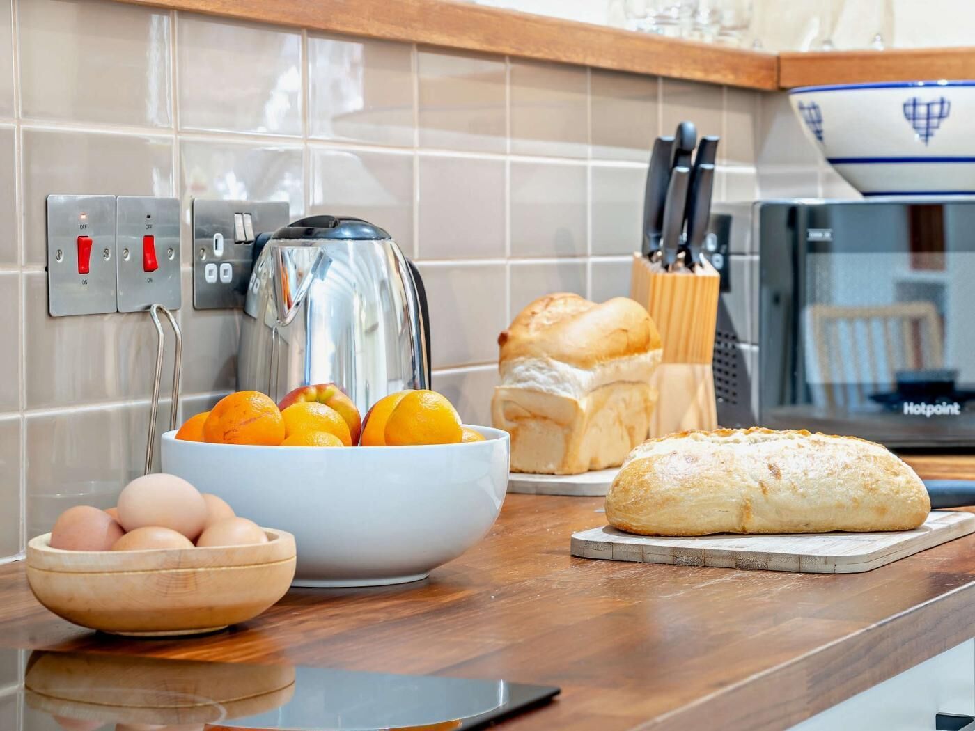 Un plan de travail de cuisine avec une bouilloire, un saladier rempli d’oranges, une miche de pain et un bol d’œufs.