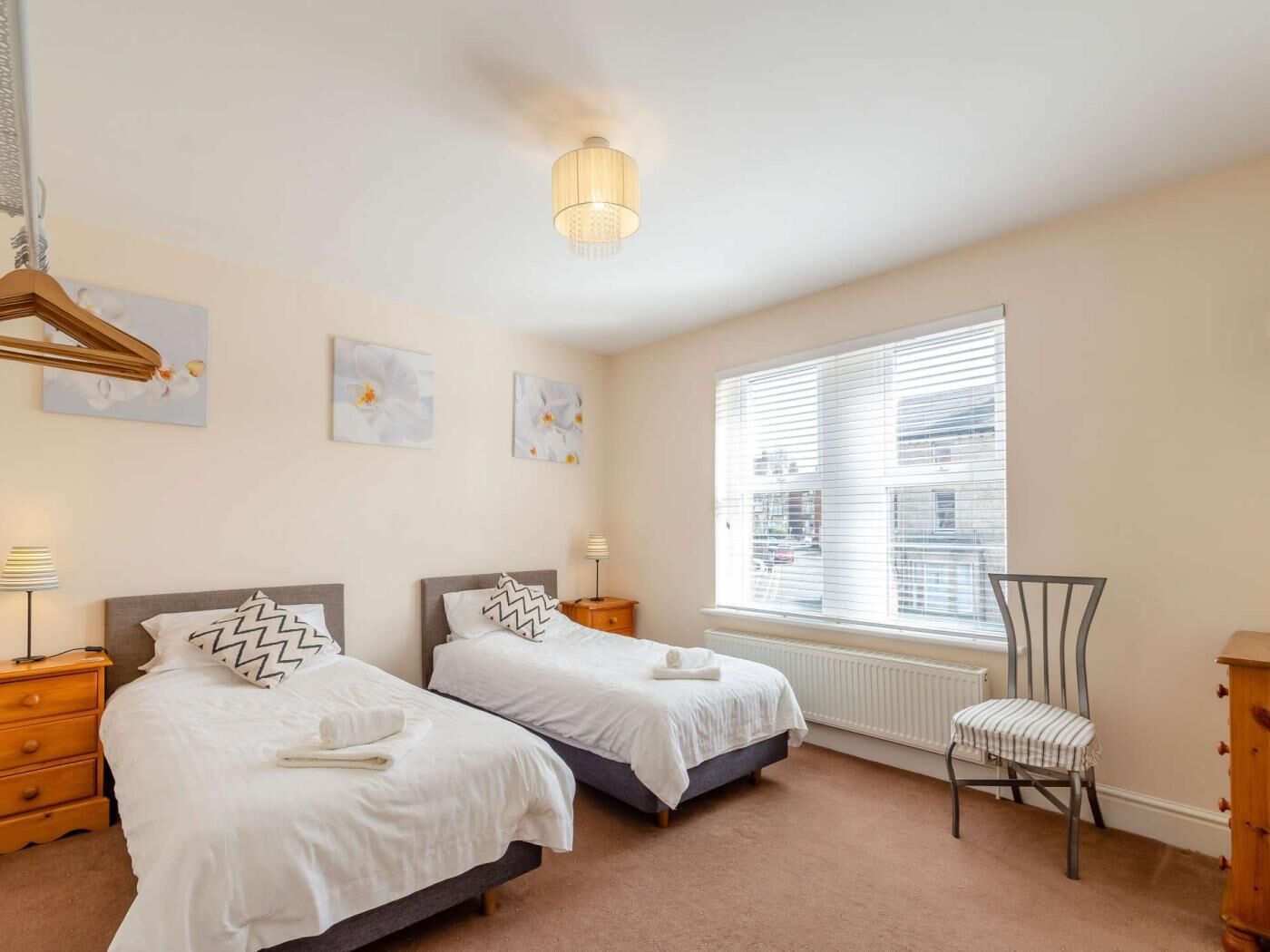 A bedroom with two beds, a radiator, a window with blinds, a chair, and a hanging light fixture.