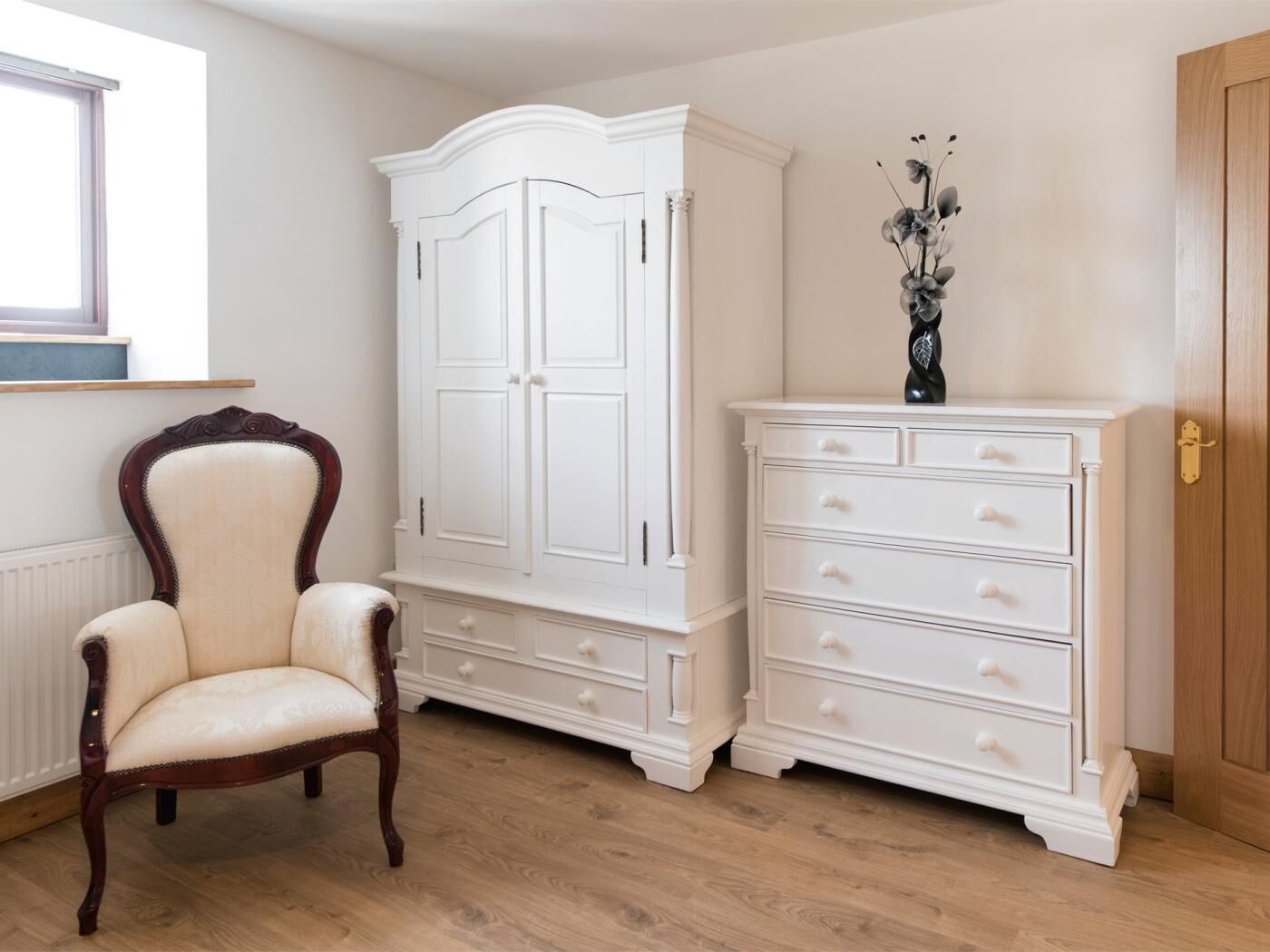 Ein Zimmer mit Holzboden, einem weißen Kleiderschrank mit Schubladen, einem weißen Schrank, einem Holzstuhl und einer Kommode mit Blumen in einer Vase.