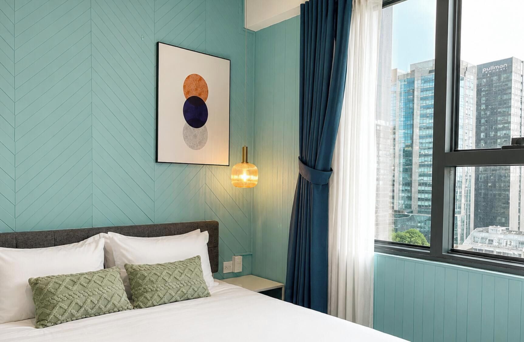 A hotel room with a bed, a wall-mounted artwork, a window with curtains, and a city view.