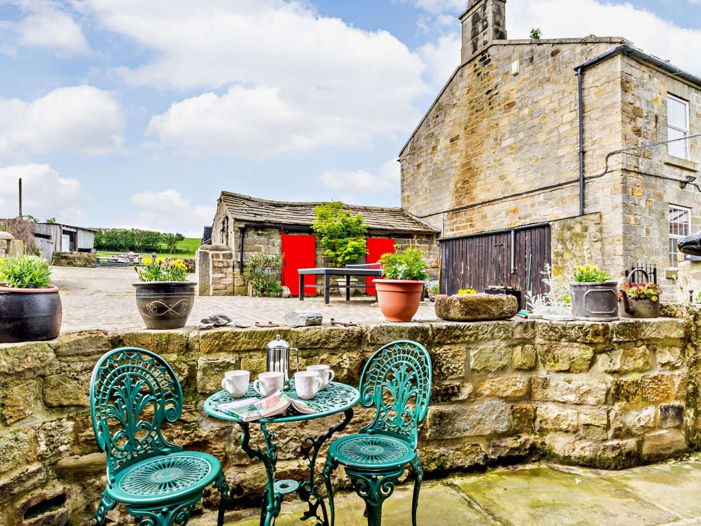 A stone building with a red door, outdoor seating with metal chairs and a table, and potted plants.