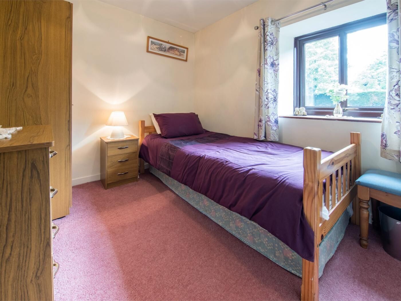 A bedroom with a bed, a bedside table with a lamp, a wooden wardrobe, and a window with curtains.