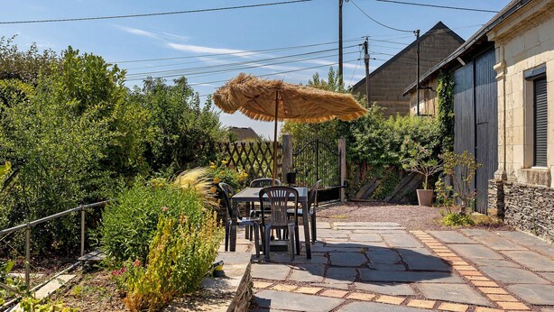 Une terrasse avec une table et des chaises, entourée de verdure et abritée par un parasol en chaume.