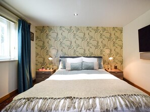 Une chambre à coucher avec un lit, deux lampes de chevet, une fenêtre avec des rideaux et un mur orné d’un motif floral.