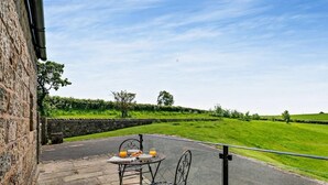 Un patio con una mesa preparada para el desayuno, un muro de piedra y vistas a un campo verde.