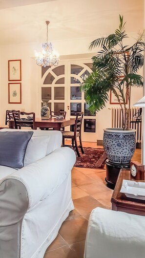 A living room with a white sofa, dining table, chairs, a chandelier, a large potted plant, and a decorative vase.