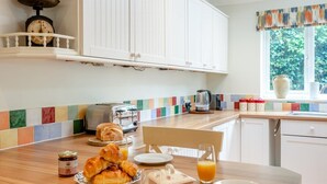 A breakfast setup with croissants, juice, and a coffee maker on a wooden table in a kitchen with white cabinets and a colorful backsplash. - 4 Bed in Old Hunstanton (oc-kt169) (Hunstanton)