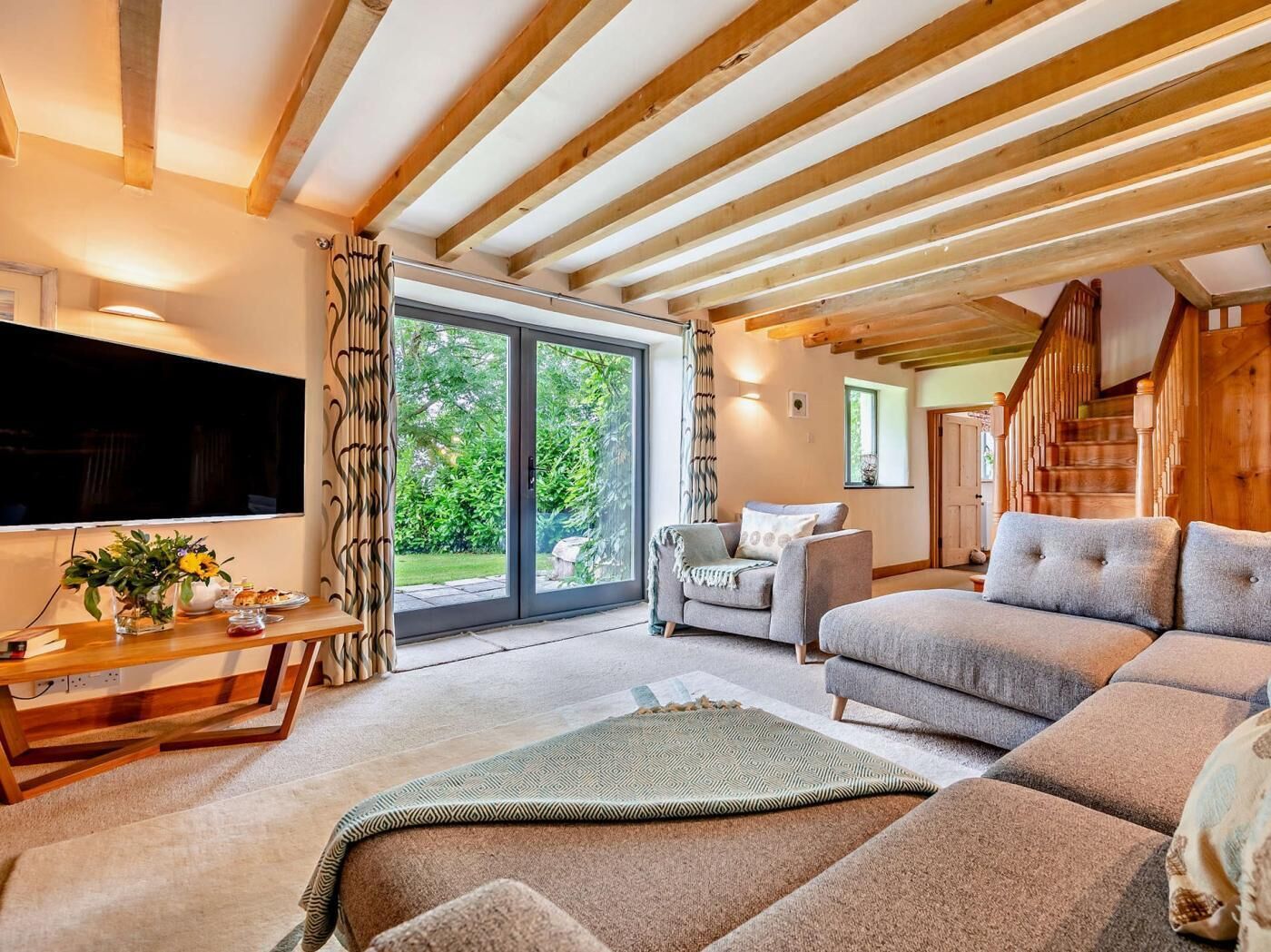 A spacious living room with a large TV, wooden ceiling beams, a sofa, a coffee table, and a sliding glass door leading to a garden.
