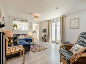 A spacious living room with a sofa, a coffee table, and a wicker chair. There is a television on a stand and a door leading to an outdoor area. - 1 Bed in Delabole (oc-65682) (Trewalder)