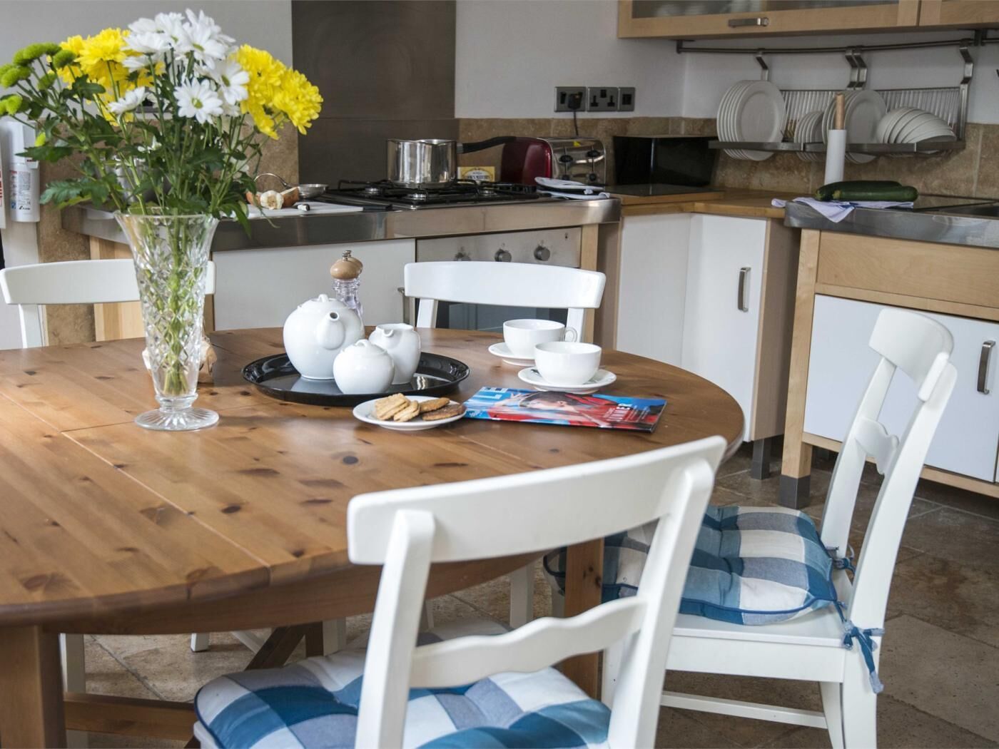 A dining area with a wooden table, white chairs, a vase of flowers, teapots, cups, and a magazine.