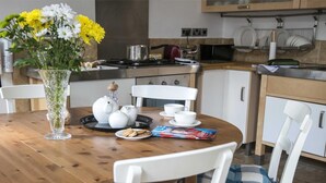 Un coin repas avec une table en bois, des chaises blanches, un vase de fleurs, des théières, des tasses et un magazine.
