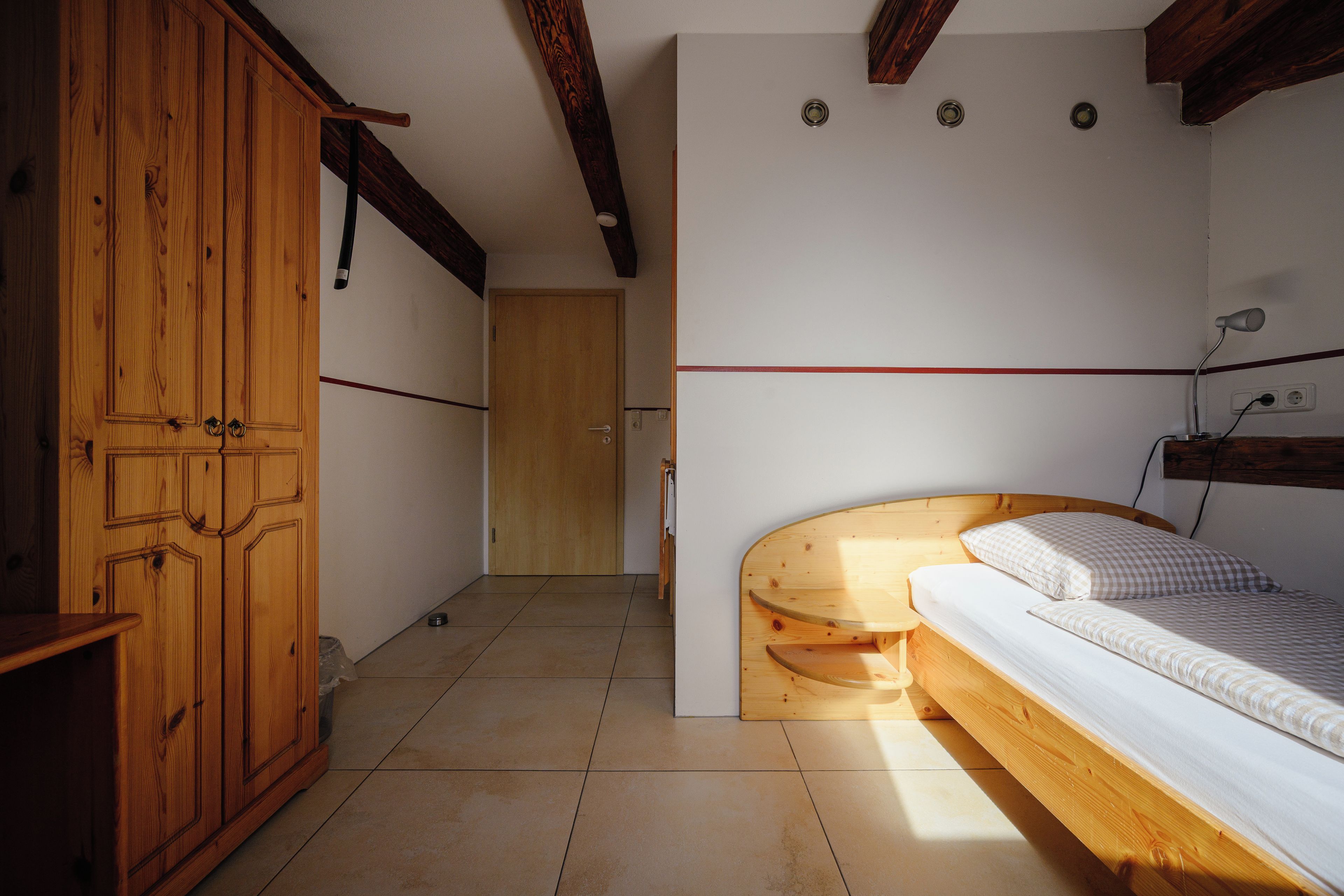 A room with a wooden wardrobe, a bed with a headboard, and a door leading to another room.