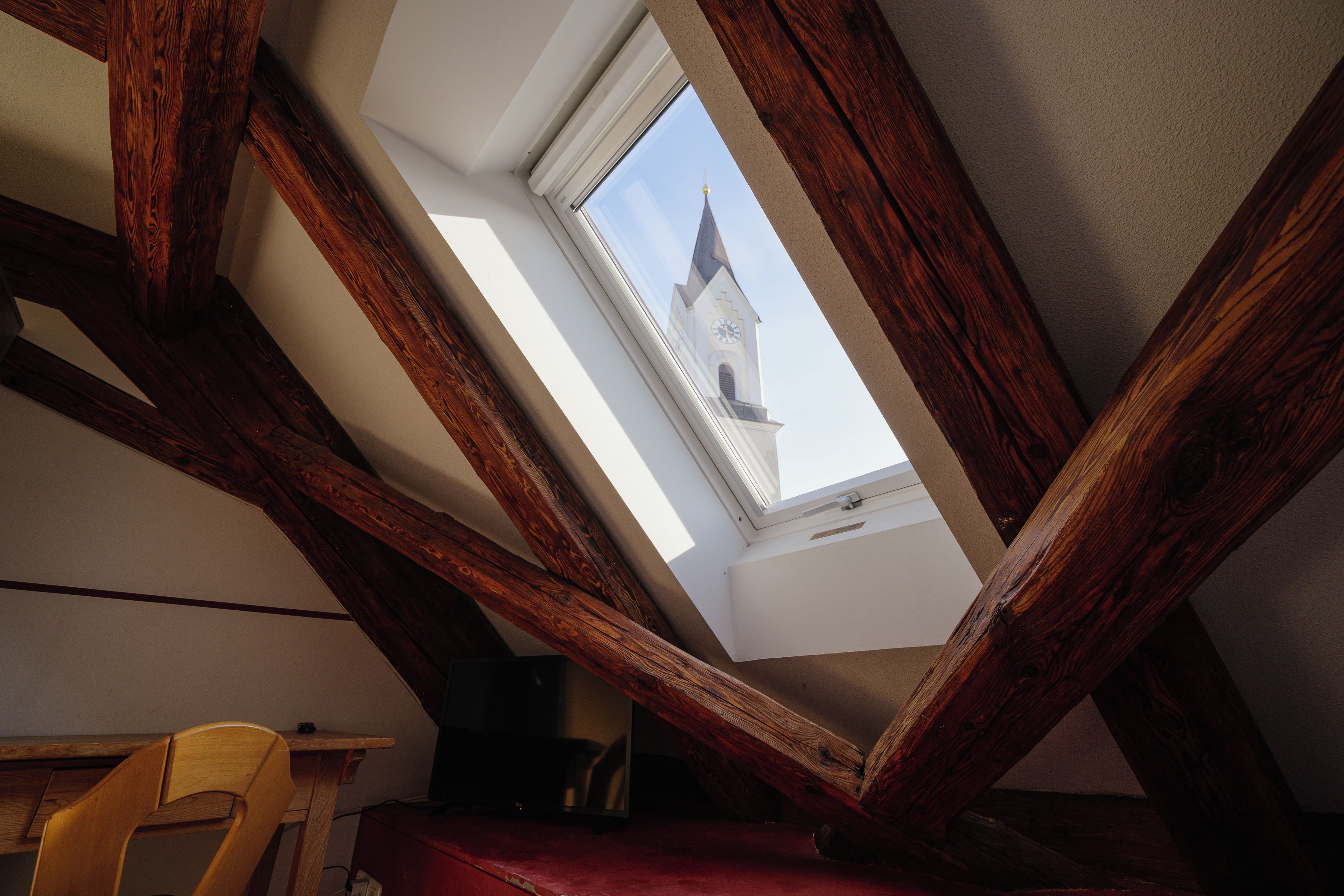 Ein Zimmer mit schrägen Wänden, Holzbalken, einem Oberlicht und Blick auf einen Kirchturm.