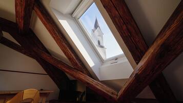 Ein Zimmer mit schrägen Wänden, Holzbalken, einem Oberlicht und Blick auf einen Kirchturm.