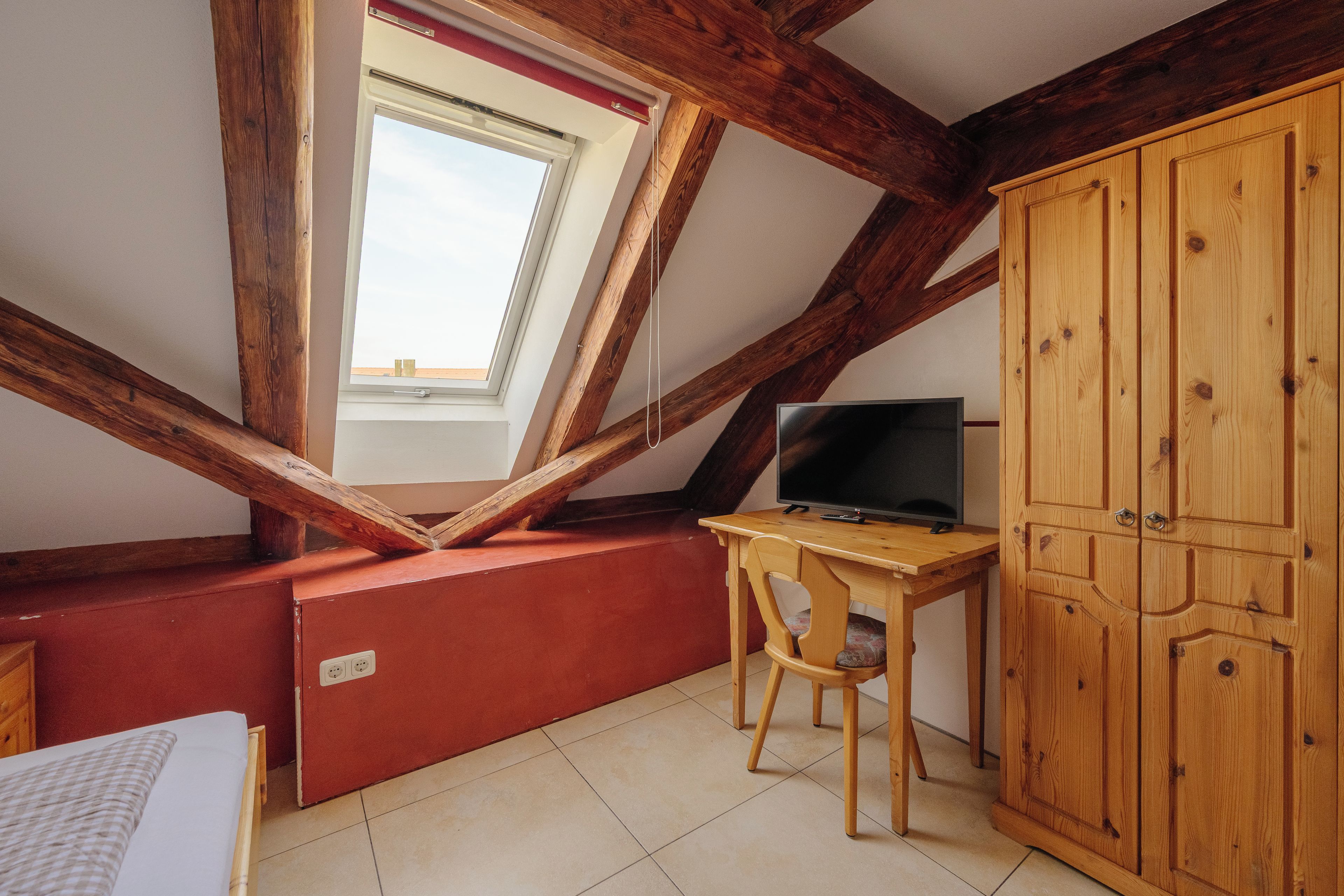 A room with a sloped ceiling, wooden beams, a television on a wooden table, a red cabinet, and a window with a view.