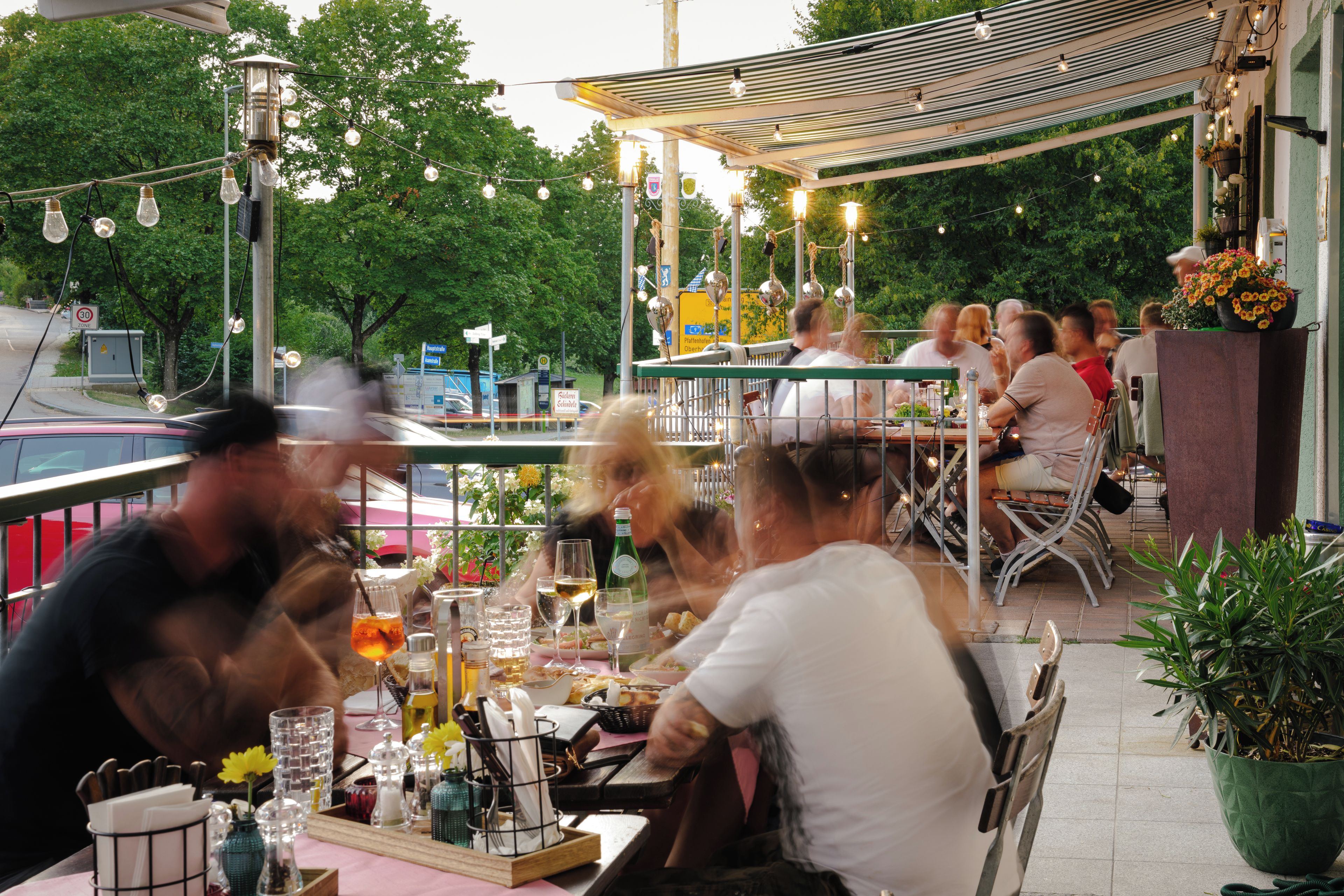 An outdoor dining area with tables and chairs, surrounded by greenery and string lights.