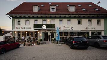 A building with multiple storefronts, including Zum Spitzbuam, Toni's, Bistro Pension, and Metzgerei. There are outdoor seating areas with tables and chairs, and cars parked in front.