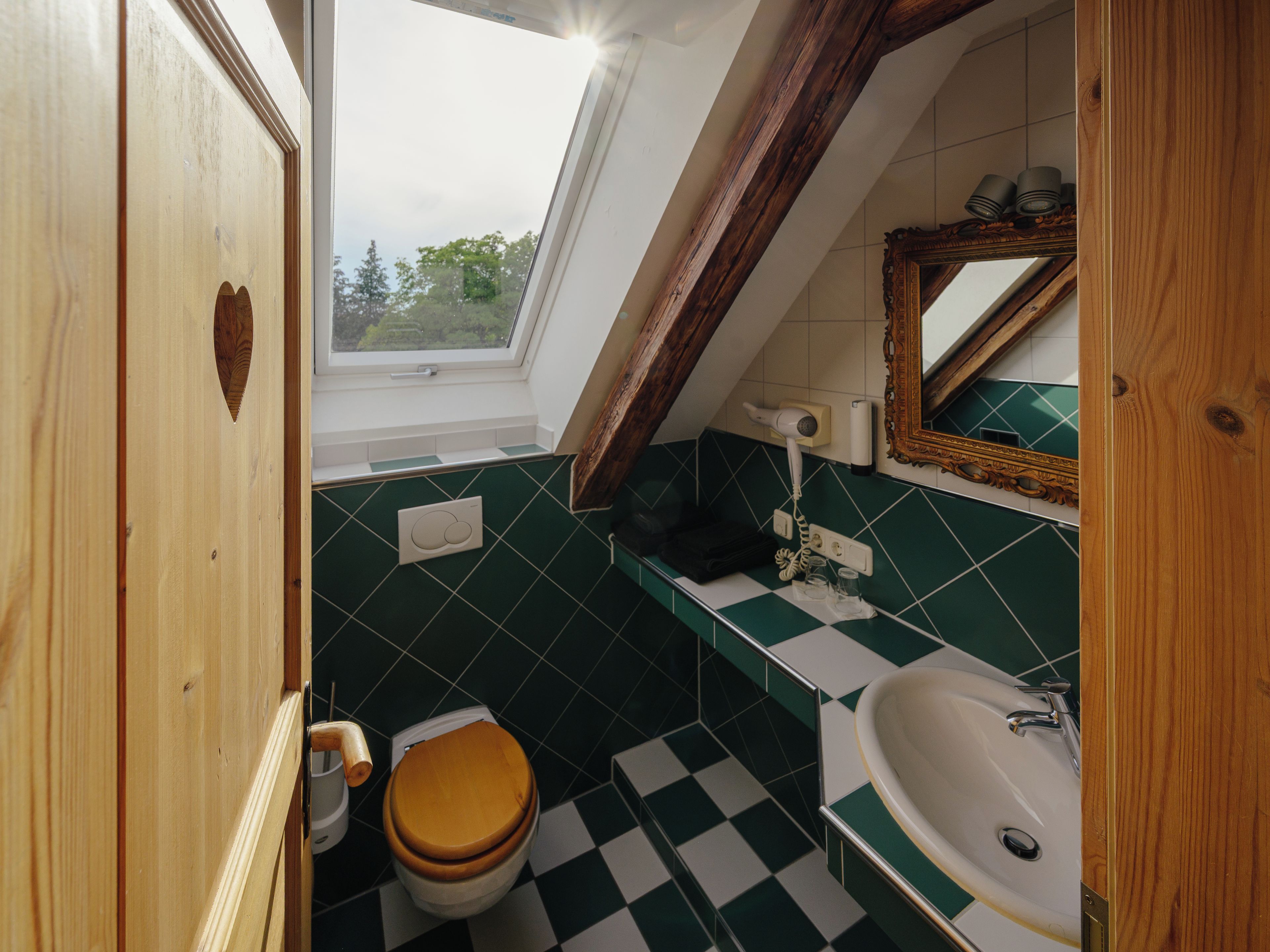 Et badeværelse med et skakternet gulv, en trædør, et toilet, en vask, et spejl og et vindue med udsigt til træer.