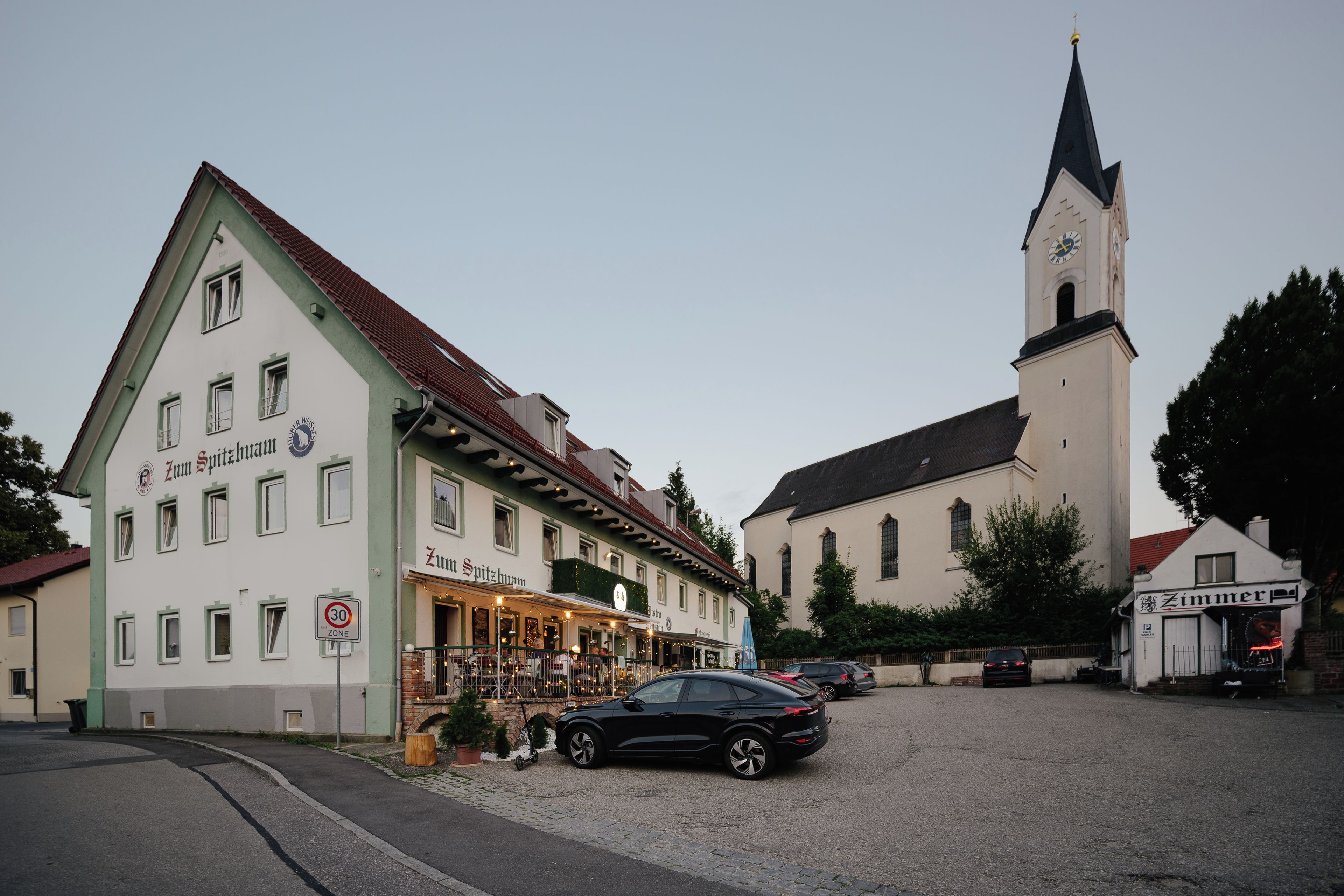 A building with a sign that reads 'Zum Spitzlhorn' and a church with a clock tower in the background.