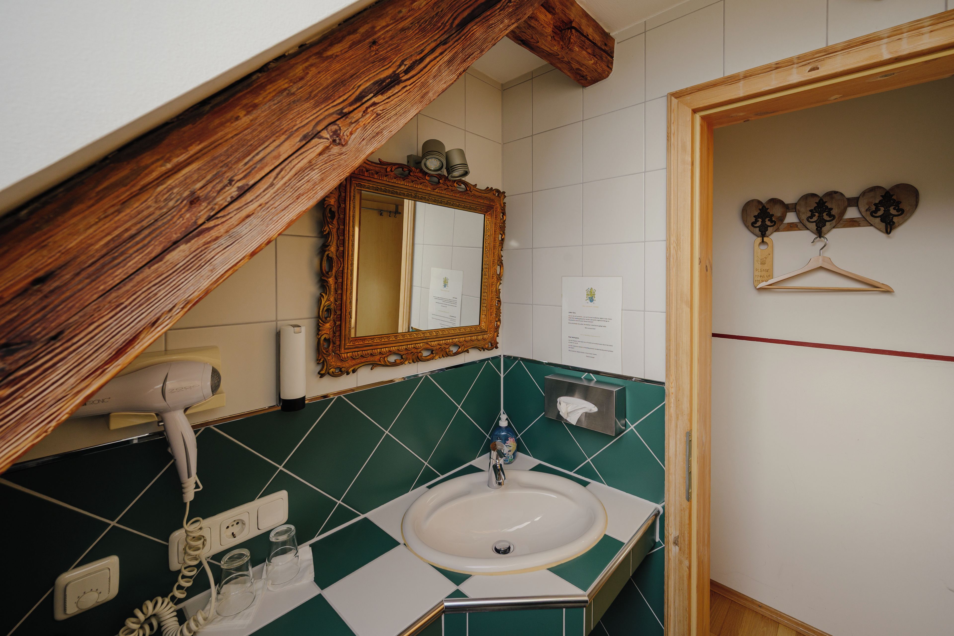 A bathroom with a white sink, green tiles, a wooden ceiling, and a mirror with a decorative frame.