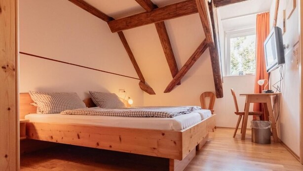 A wooden bed in a room with exposed beams, a desk with a chair, and a television on the wall.