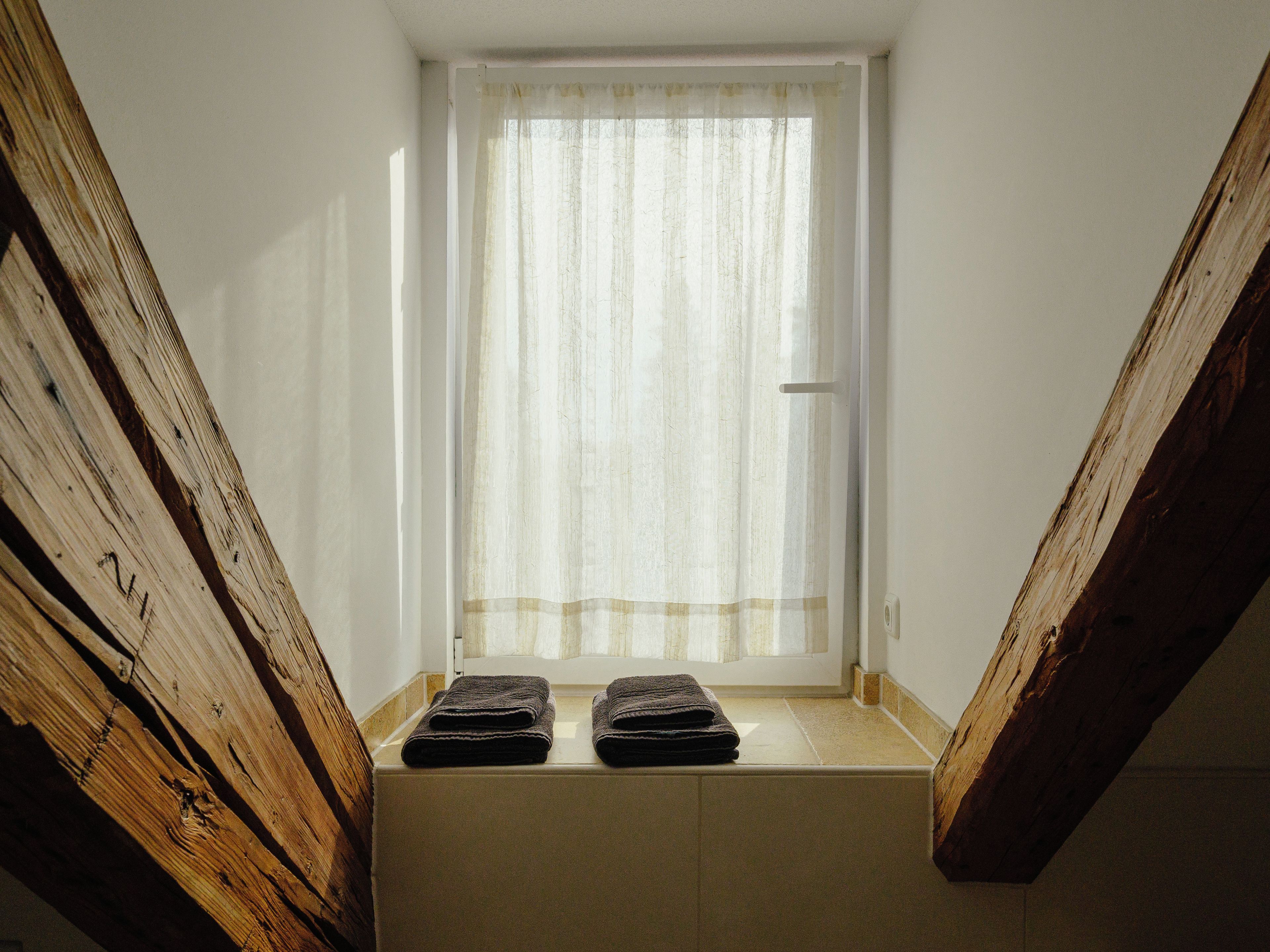 A narrow attic window with a white curtain, wooden beams, and two folded towels on the sill.