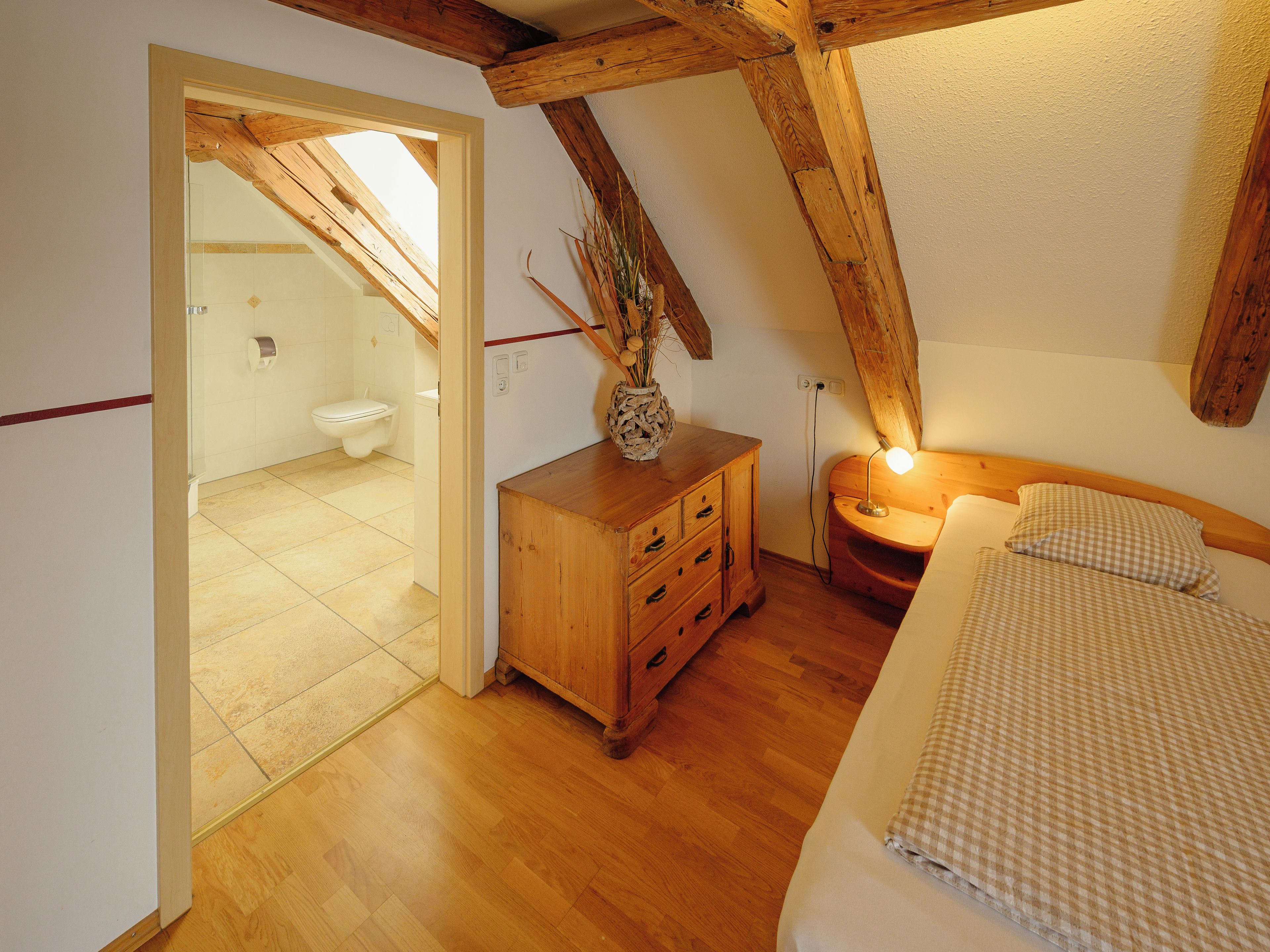 A bedroom with a wooden ceiling, a bed with checkered bedding, a bedside table with a lamp, and a wooden dresser.