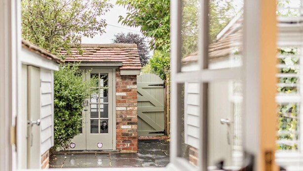 A view through a window of a house with a brick wall, a green door, and a small garden area. - 1 Bed in Tenterden (oc-66342) (Tenterden)