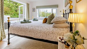 A bedroom with a large bed, a bedside table with a lamp, a window with curtains, and a view of greenery outside. - 2 Bed in Bangor (oc-66276) (Llanddeiniolen)